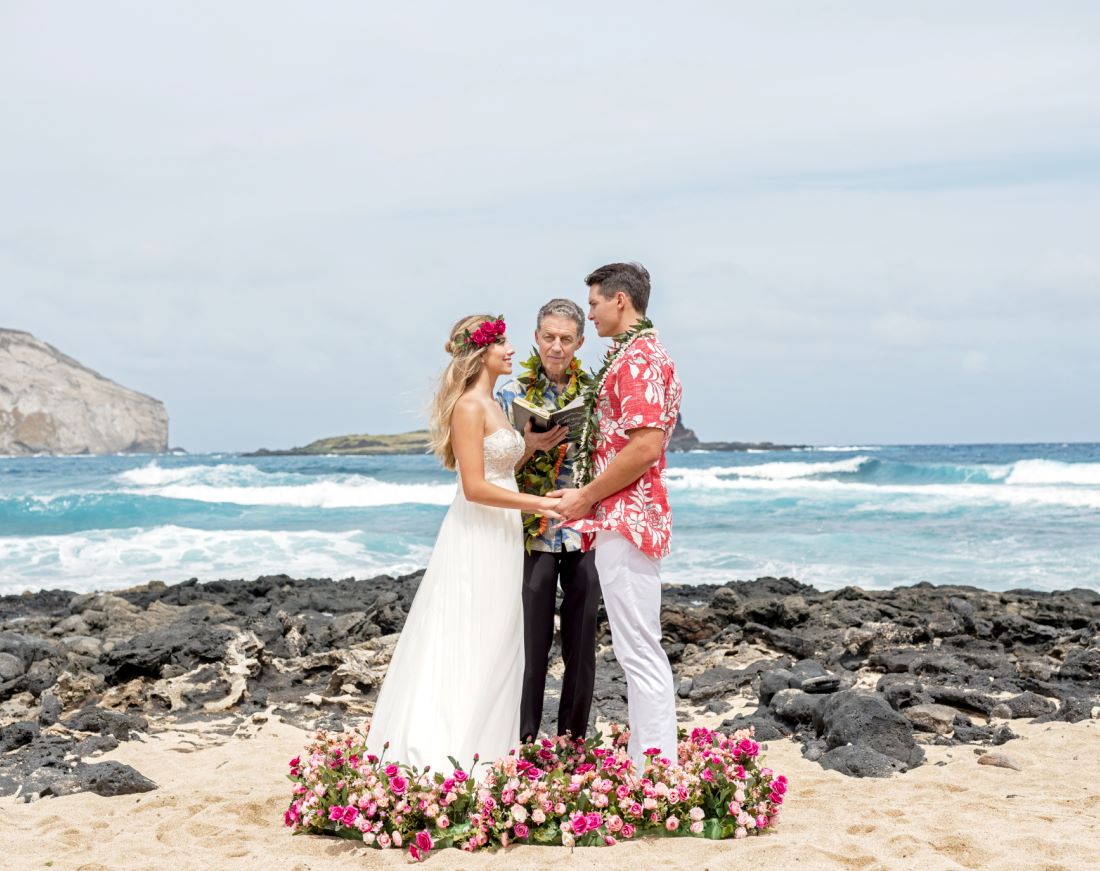 A couple exchanging vows with an officiant on the sand, a low arc of pink roses at their feet and surf behind