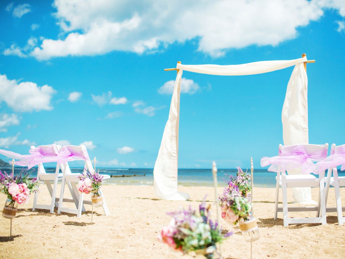 A beach ceremony set up with a draped arch, white chairs and flower arrangements on the sand