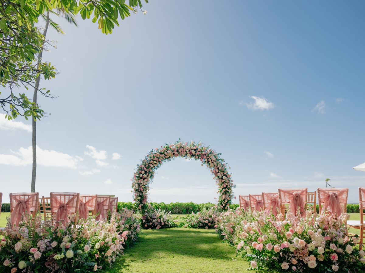A resort lawn ceremony with a full floral arch, blush chair sashes and flower-lined aisle, ocean beyond