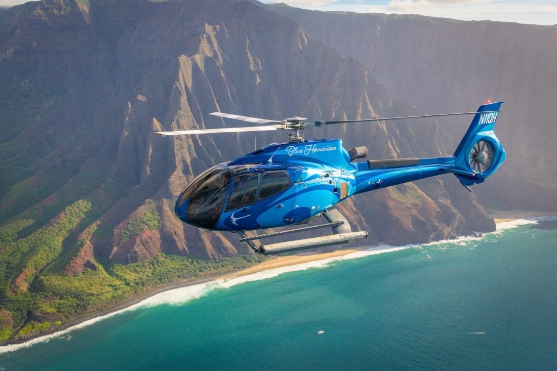 A Blue Hawaiian helicopter flying along the fluted cliffs of the Na Pali Coast above a white sand beach