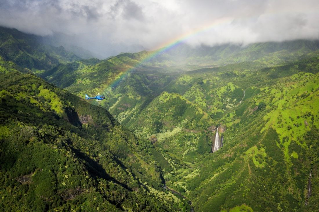 A rainbow over the green interior of Kauai with a waterfall and a Blue Hawaiian helicopter in flight