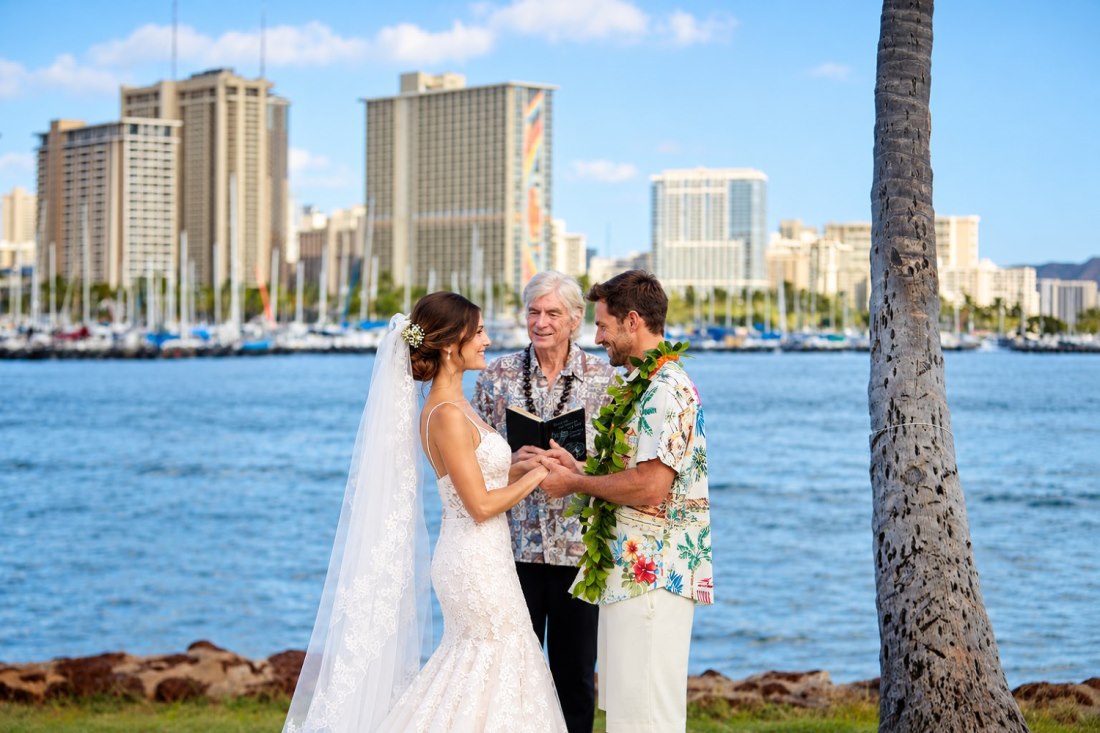 A couple exchanging vows with an officiant at Magic Island, the Waikiki skyline and yacht harbour behind