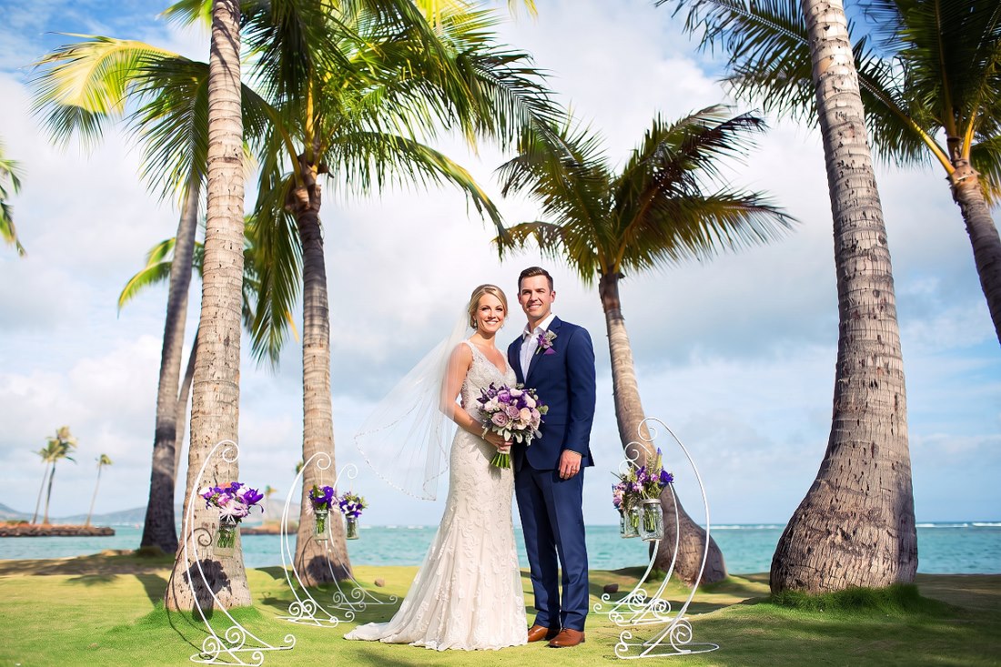 A bride and groom on a palm-shaded lawn between aisle stands holding mason jar flowers, ocean behind