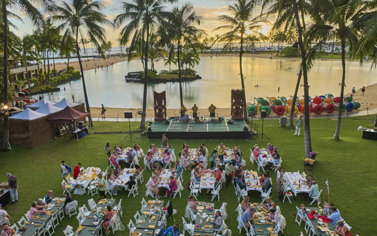 Long tables and tiki torches on the lawn beside Duke Kahanamoku Lagoon at the Waikiki Starlight Luau, with the ocean and palms behind the stage at sunset